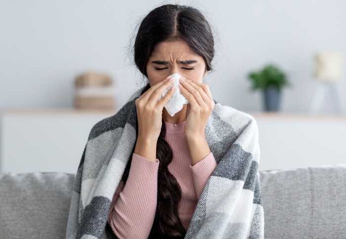 A woman covered in blanket sitting on a sofa and blowing nose
